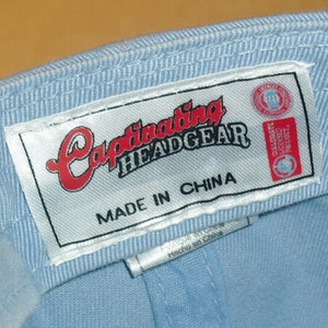 Pu&ograve; includere: Un berretto da baseball blu chiaro con un'etichetta bianca che recita "Captivating Headgear Made in China".