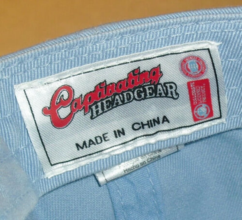 Pu&ograve; includere: Un berretto da baseball blu chiaro con un'etichetta bianca che recita "Captivating Headgear Made in China".