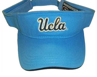 May include: A blue visor with a navy blue and gold embroidered "UCLA" logo. The visor is made by Colosseum.