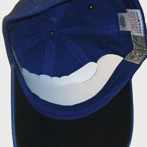 May include: Royal blue baseball cap with a black peak. The cap has the gold letters "KC" embroidered on the front. The underside of the peak is white. The cap has a velcro strap for adjustment.