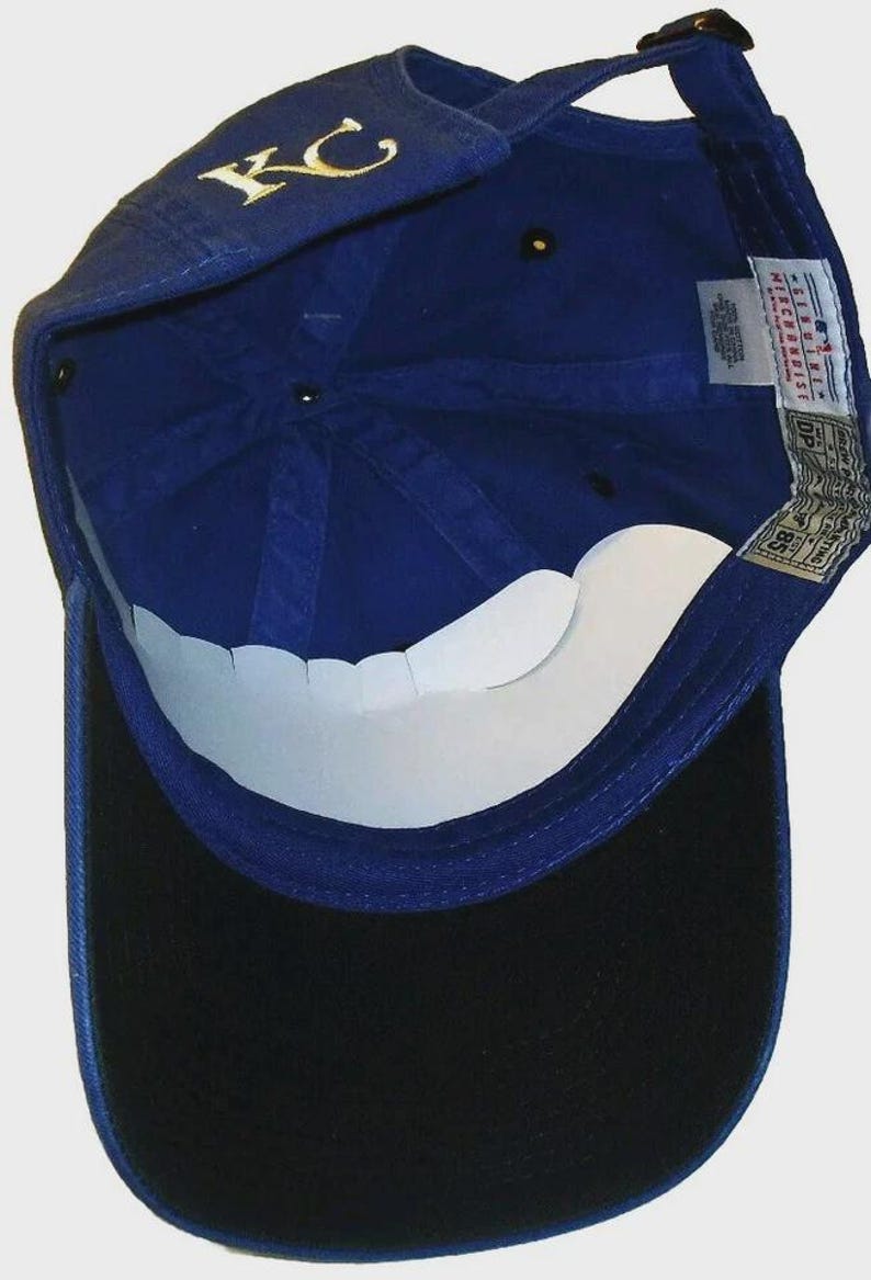 May include: Royal blue baseball cap with a black peak. The cap has the gold letters "KC" embroidered on the front. The underside of the peak is white. The cap has a velcro strap for adjustment.