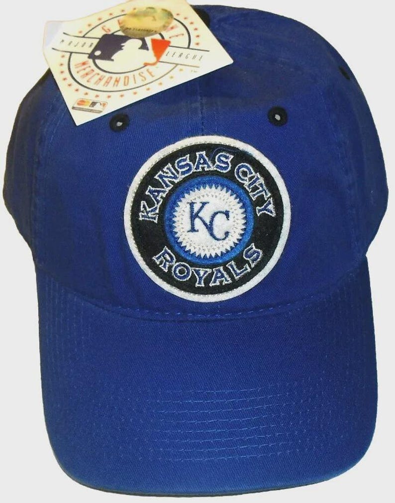 May include: A royal blue baseball cap with the Kansas City Royals logo. The logo is a circular patch with the team name and "KC" in the centre. A Major League Baseball merchandise tag is attached.