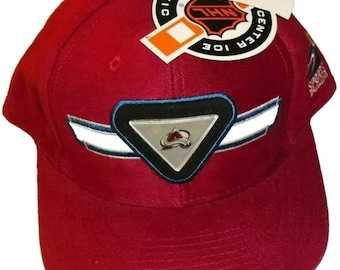 May include: A red baseball cap with a white and blue logo of the Colorado Avalanche hockey team. The cap has a white stripe with a blue trim and a silver triangle with the team's logo. The cap has a tag attached that says "Sports Specialties" and "Center Ice".