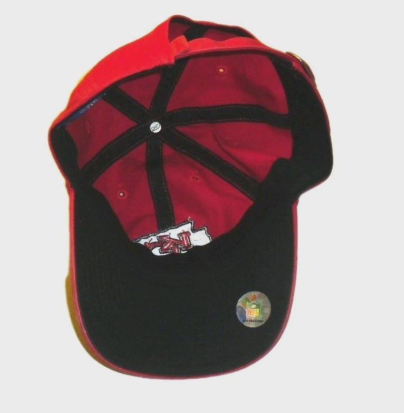 May include: A red baseball cap viewed from the inside, featuring black interior seam tape, a black brim underside, and a white embroidered logo patch. A circular NFL holographic sticker is affixed to the black brim, and a small Reebok logo tag is visible on the interior crown.