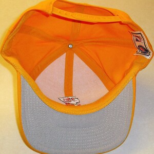 May include: A yellow baseball cap with a gray brim. The inside of the cap is yellow and has a white label with red text. The back of the cap has a white label with a red logo.