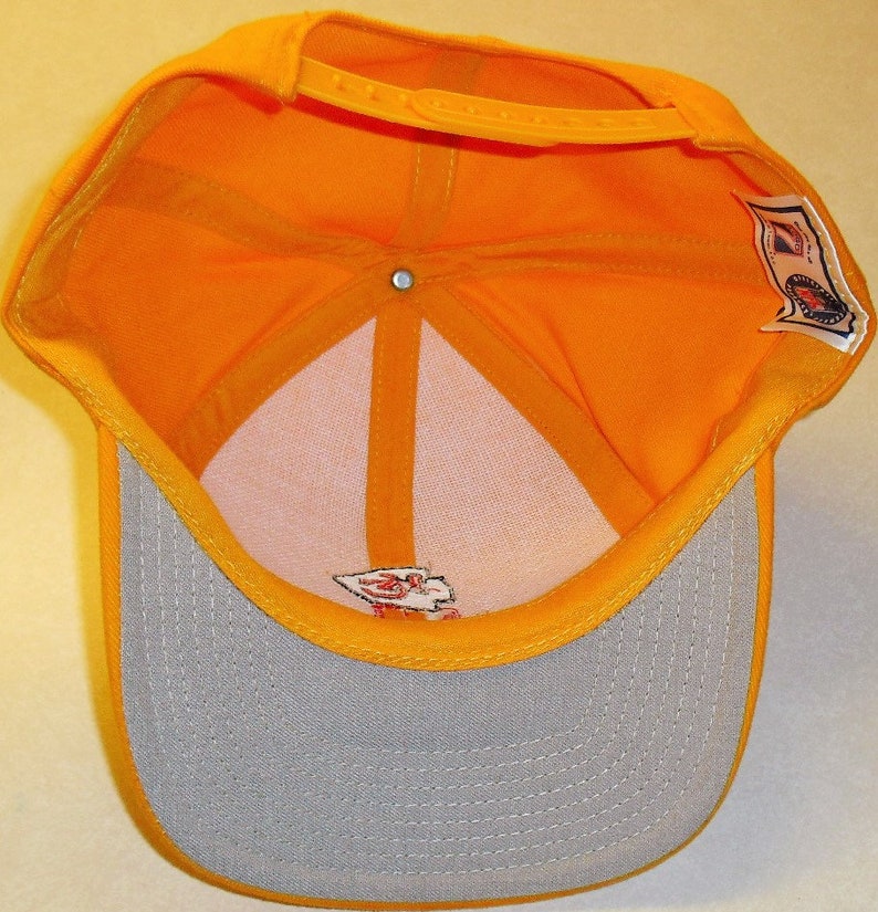 May include: A yellow baseball cap with a gray brim. The inside of the cap is yellow and has a white label with red text. The back of the cap has a white label with a red logo.