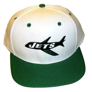 May include: White baseball cap with a green bill. The front of the cap features a green airplane with the word "JETS" in black and white lettering.