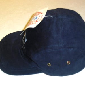 K&ouml;nnte beinhalten: Eine marineblaue Baseballkappe mit einem gebogenen Schirm und einer flachen Oberseite. Die Kappe hat ein kleines Etikett an der Seite.