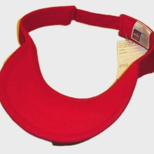 May include: A bright red visor with a curved brim and adjustable strap. The visor has a tag attached with text. The visor is designed to shield the eyes from the sun.