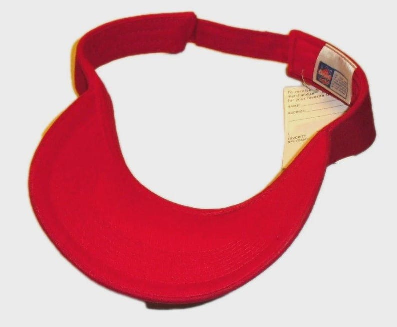 May include: A bright red visor with a curved brim and adjustable strap. The visor has a tag attached with text. The visor is designed to shield the eyes from the sun.