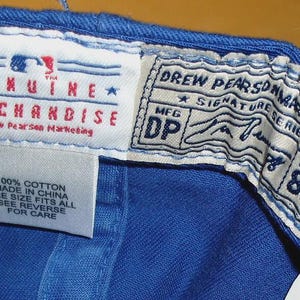 May include: Detail of the inside of a blue baseball cap, showing multiple tags. The tags display text such as "GENUINE MERCHANDISE" and "DREW PEARSON MARKETING". The labels also state the cap is 100% cotton and one size fits all.