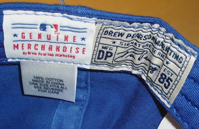 May include: Detail of the inside of a blue baseball cap, showing multiple tags. The tags display text such as "GENUINE MERCHANDISE" and "DREW PEARSON MARKETING". The labels also state the cap is 100% cotton and one size fits all.