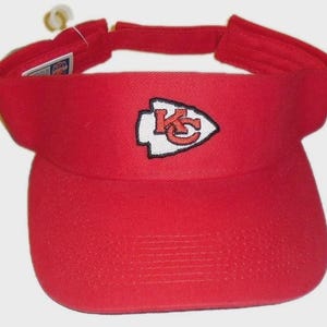 May include: A red visor featuring the Kansas City Chiefs logo. The logo is a white arrowhead with a red "KC" inside. The visor has a curved brim and an adjustable strap. The visor is made of a durable fabric.
