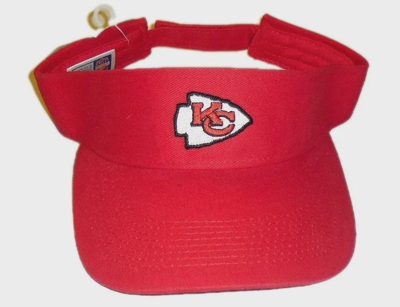 May include: A red visor featuring the Kansas City Chiefs logo. The logo is a white arrowhead with a red "KC" inside. The visor has a curved brim and an adjustable strap. The visor is made of a durable fabric.