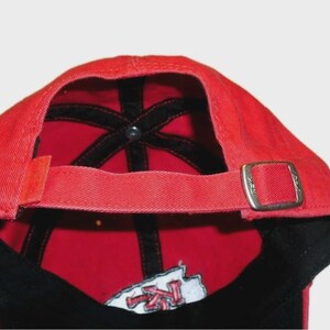 May include: A red baseball cap viewed from the back, featuring a fabric strap with a silver-toned metal sliding buckle for adjustment. The interior of the cap shows black seam tape and a partial embroidered patch with red and white lettering.