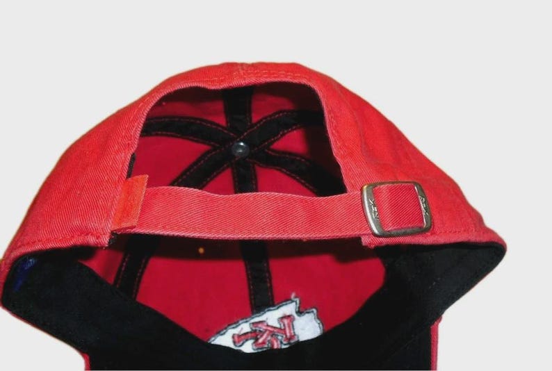 May include: A red baseball cap viewed from the back, featuring a fabric strap with a silver-toned metal sliding buckle for adjustment. The interior of the cap shows black seam tape and a partial embroidered patch with red and white lettering.