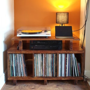 May include: A wooden record player stand with three shelves. The top shelf has a turntable, a laptop, and a lamp with a black shade. The middle shelf has a black stereo receiver. The bottom shelf has vinyl records.
