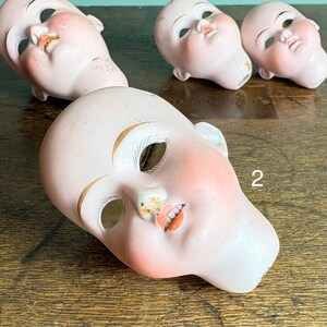 May include: Four antique porcelain doll heads. The heads are painted with rosy cheeks, open mouths, and detailed facial features. The eyes are missing. The number "2" is visible on one head. The heads are on a wooden surface.