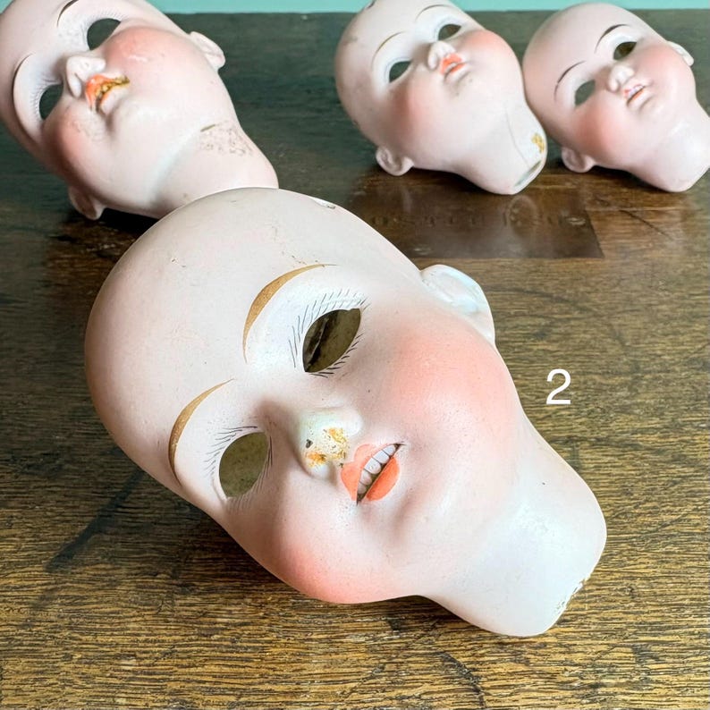 May include: Four antique porcelain doll heads. The heads are painted with rosy cheeks, open mouths, and detailed facial features. The eyes are missing. The number "2" is visible on one head. The heads are on a wooden surface.
