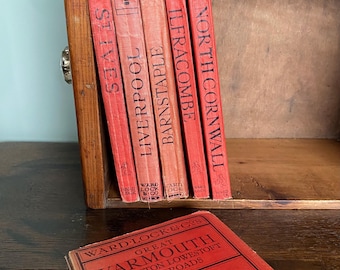 May include: A collection of vintage red books with titles including "St Ives," "Liverpool," and "North Cornwall." The books are arranged in a wooden shelf, with one book lying in front. The books are from Ward Lock & Co.