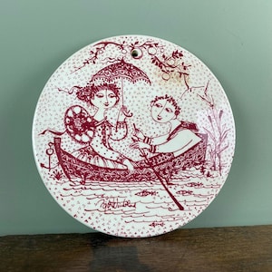 Pode incluir: Um prato de cerâmica branco com um desenho vermelho de um casal em um barco com um guarda-chuva. O prato é decorado com um padrão vermelho e branco e tem uma assinatura na parte inferior.