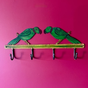Puede incluir: Un colgador de llaves decorativo montado en la pared con dos figuras de loro verde posadas en una barra de madera verde. Los loros tienen detalles rojos y se miran. Cuatro ganchos negros cuelgan debajo de la barra, sobre un fondo magenta.
