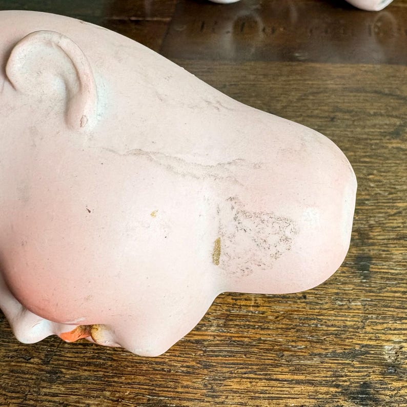 May include: A close-up of a vintage porcelain doll head. The head is painted a pale pink with visible wear and tear, including discoloration and small imperfections. The ear is visible, and the doll's face is partially visible.