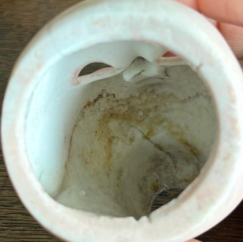 May include: A close-up view of the inside of a white porcelain doll head. The interior shows a hollow space with a crack and discoloration. The rim of the head is a soft pink color.