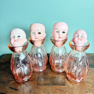 May include: Four antique glass bottles with doll heads. The bottles are a light pink color with a textured, teardrop design. Each bottle has a copper-colored collar and a porcelain doll head. The doll heads have painted features and are in various states of disrepair.