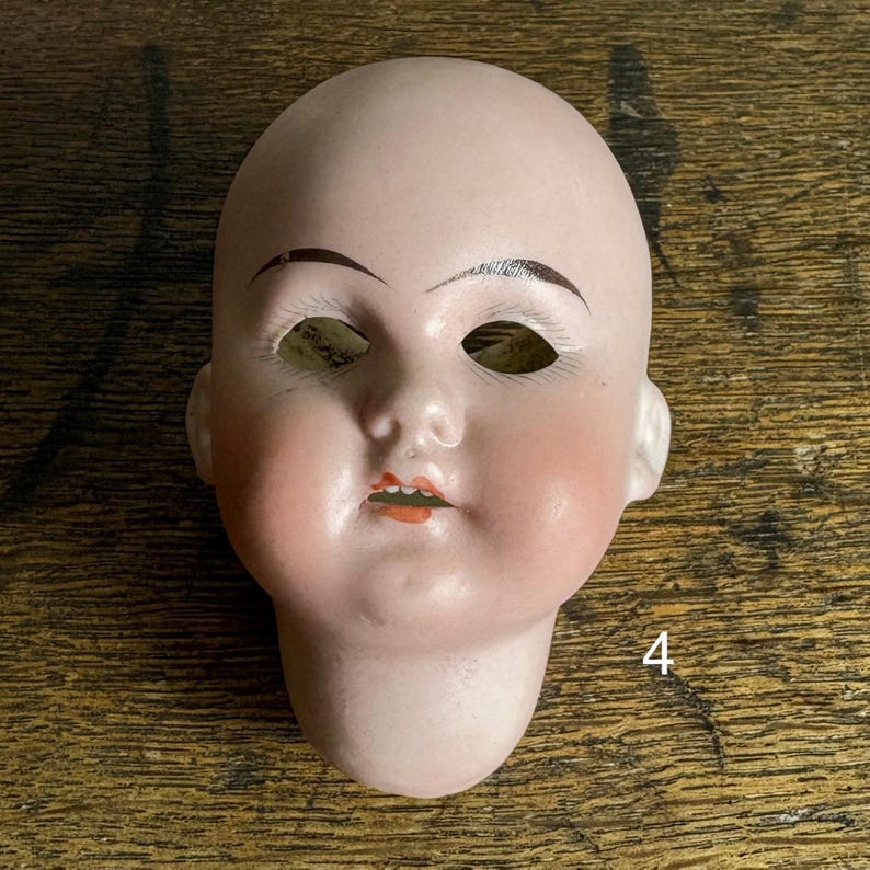 May include: A vintage doll head with a pale complexion, painted eyebrows, and rosy cheeks. The doll head has open eye sockets and a small, open mouth with painted lips. The number "4" is visible on the lower right side.