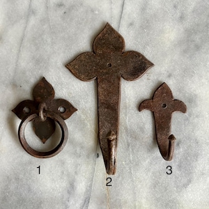 Może przedstawiać: Trzy metalowe haczyki w stylu antycznym i uchwyt pierścieniowy. Haczyki mają wzór fleur-de-lis i rdzawobrązowe wykończenie. Uchwyt pierścieniowy jest okrągły i przymocowany do ozdobnej płyty. Przedmioty są prawdopodobnie przeznaczone do wieszania przedmiotów.