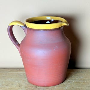 May include: A red terracotta pitcher with a yellow rim and a brown glaze. The pitcher has a rounded body and a handle.