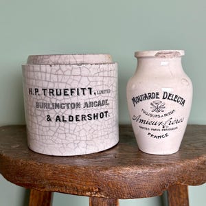 May include: Two vintage ceramic jars on a wooden surface. The jar on the left is cylindrical with the text "H.P. TRUEFITT, LIMITED BURLINGTON ARCADE, & ALDERSHOT." The jar on the right is a vase shape with the text "MOUTARDE DELECTA Amieux frères FRANCE."