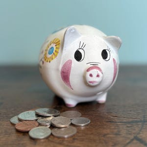 May include: A white ceramic piggy bank with painted details, including pink cheeks, black eyes, and floral accents. A small pile of coins rests in front of the bank on a wooden surface. The piggy bank is a classic design.