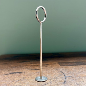 May include: A silver-colored place card holder with a circular top and a round base. The holder has a slender, straight stem. The background is a muted green wall and a wooden surface.