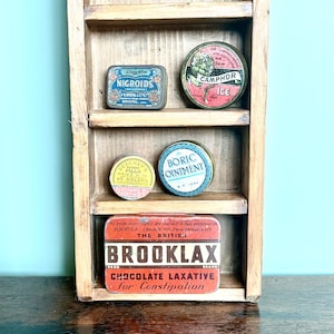 Può includere: Una mensola in legno espone barattoli e contenitori in metallo vintage. Gli oggetti includono una scatola rettangolare blu "Nigroids", una scatola rotonda "Camphor Ice", una scatola rotonda "Boric Ointment", una scatola rotonda "Beecham's Pills" e una scatola rettangolare "Brooklax".