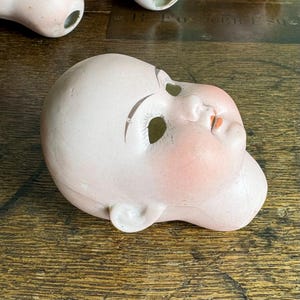 May include: A vintage porcelain doll head with painted features, including rosy cheeks, brown eyebrows, and red lips. The doll head is a pale pink color and has eye sockets and ear details. The head is lying on a wooden surface.
