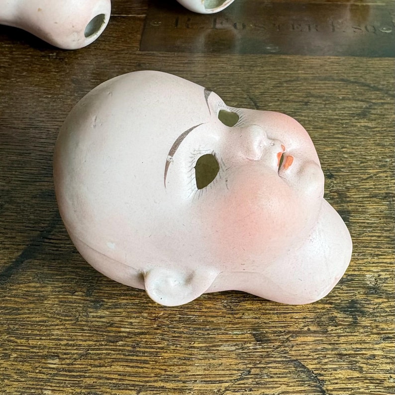May include: A vintage porcelain doll head with painted features, including rosy cheeks, brown eyebrows, and red lips. The doll head is a pale pink color and has eye sockets and ear details. The head is lying on a wooden surface.