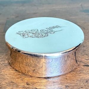 May include: A small, round silver box with a light blue lid. The lid features an embossed floral design. The box has a shiny, reflective surface and sits on a wooden surface. The box is a decorative item.