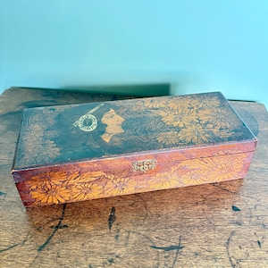 May include: A rectangular wooden box with a hinged lid and a brass latch. The surface features a dark brown wood grain finish with an engraved illustration of a profile of a person surrounded by detailed floral patterns on the lid and front.