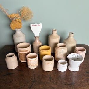 May include: A collection of vintage ceramic jars and bottles in various sizes and shapes. The jars are in shades of beige and cream, with some having a textured surface. The bottles are also beige and cream, with some having a clear glass finish. The jars and bottles are arranged on a wooden surface. A small white sign with the text "Hartley & Lord" is visible in the background.