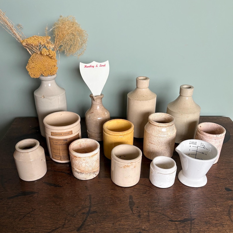 May include: A collection of vintage ceramic jars and bottles in various sizes and shapes. The jars are in shades of beige and cream, with some having a textured surface. The bottles are also beige and cream, with some having a clear glass finish. The jars and bottles are arranged on a wooden surface. A small white sign with the text "Hartley & Lord" is visible in the background.