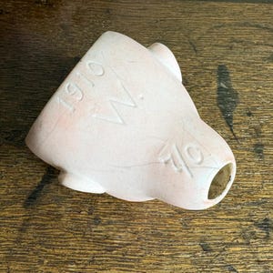 May include: A light pink ceramic object with the numbers "1910" and "40" and the letter "W" etched into the surface. The object has a rounded shape with a hole at one end.