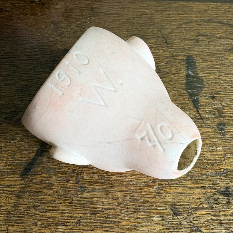 May include: A light pink ceramic object with the numbers "1910" and "40" and the letter "W" etched into the surface. The object has a rounded shape with a hole at one end.