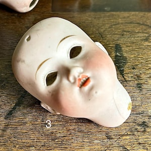 May include: A vintage porcelain doll head with painted facial features. The doll head has open eye sockets, a small mouth, and rosy cheeks. The number "3" is printed on the doll's chin. The head is off-white with gold accents.
