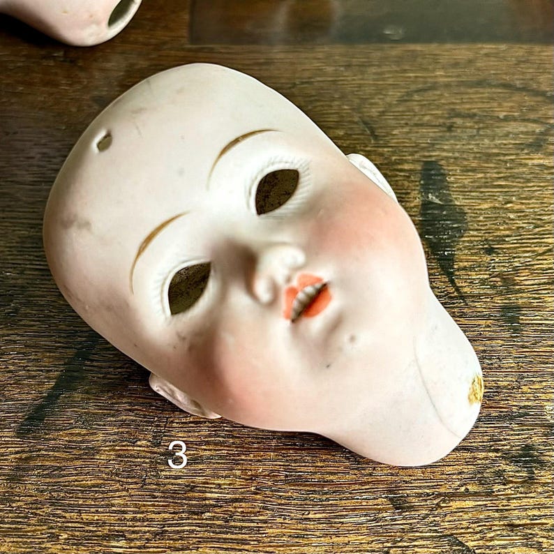 May include: A vintage porcelain doll head with painted facial features. The doll head has open eye sockets, a small mouth, and rosy cheeks. The number "3" is printed on the doll's chin. The head is off-white with gold accents.