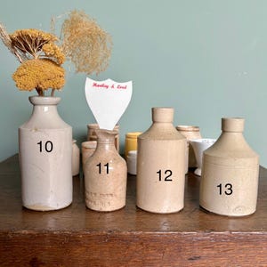 May include: Five vintage stoneware bottles with cork tops. The bottles are numbered 10, 11, 12, and 13. The bottles are all different sizes and shapes. The bottles are on a wooden table.