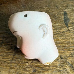 May include: A vintage porcelain doll head, featuring a pale complexion with delicate blush on the cheeks and lips. The head has closed eyes, a detailed ear, and a small hole at the top. The porcelain has a smooth finish, with slight wear.