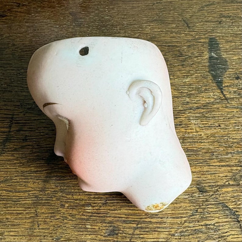 May include: A vintage porcelain doll head, featuring a pale complexion with delicate blush on the cheeks and lips. The head has closed eyes, a detailed ear, and a small hole at the top. The porcelain has a smooth finish, with slight wear.