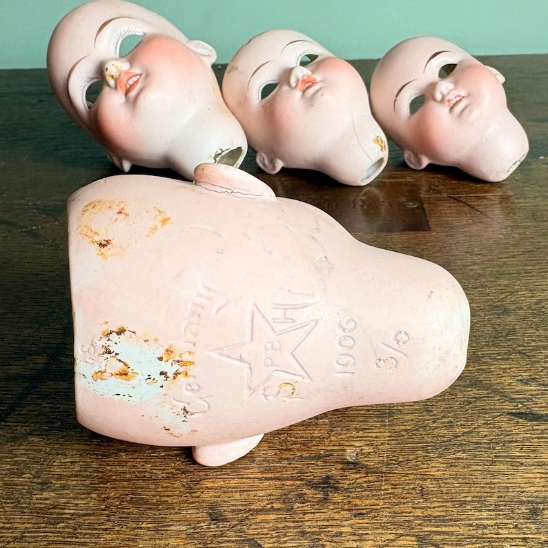 May include: Four antique doll parts: three porcelain doll heads and a pink torso. The doll heads are painted with delicate features, including closed eyes and rosy cheeks. The torso is marked with text and a star. The items are arranged on a wooden surface.